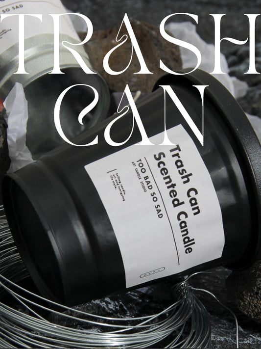 Black candle labeled 'Trash Can Scented Candle' with a label reading 'Too Bad So Sad' on a dark background.