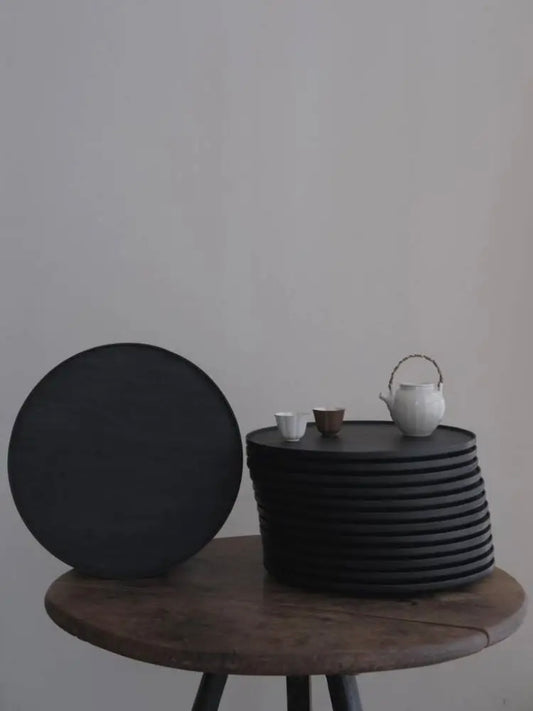 Black round stool and textured black side table with a white teapot and cups on a plain background