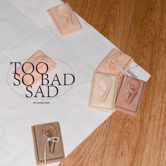 Artisanal candles shaped like ears on a wooden surface with 'Too So Bad Sad' branding.