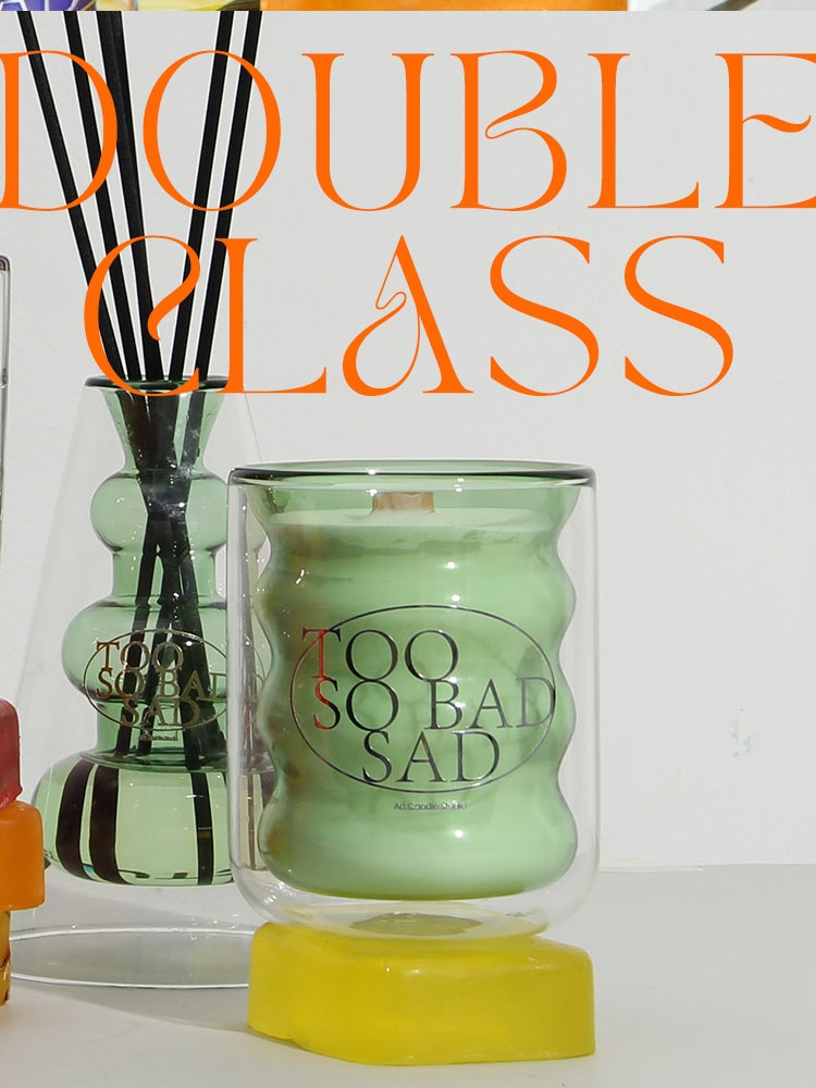 Green candle with 'Too Sad' text and diffuser sticks in a clear container on a white background.