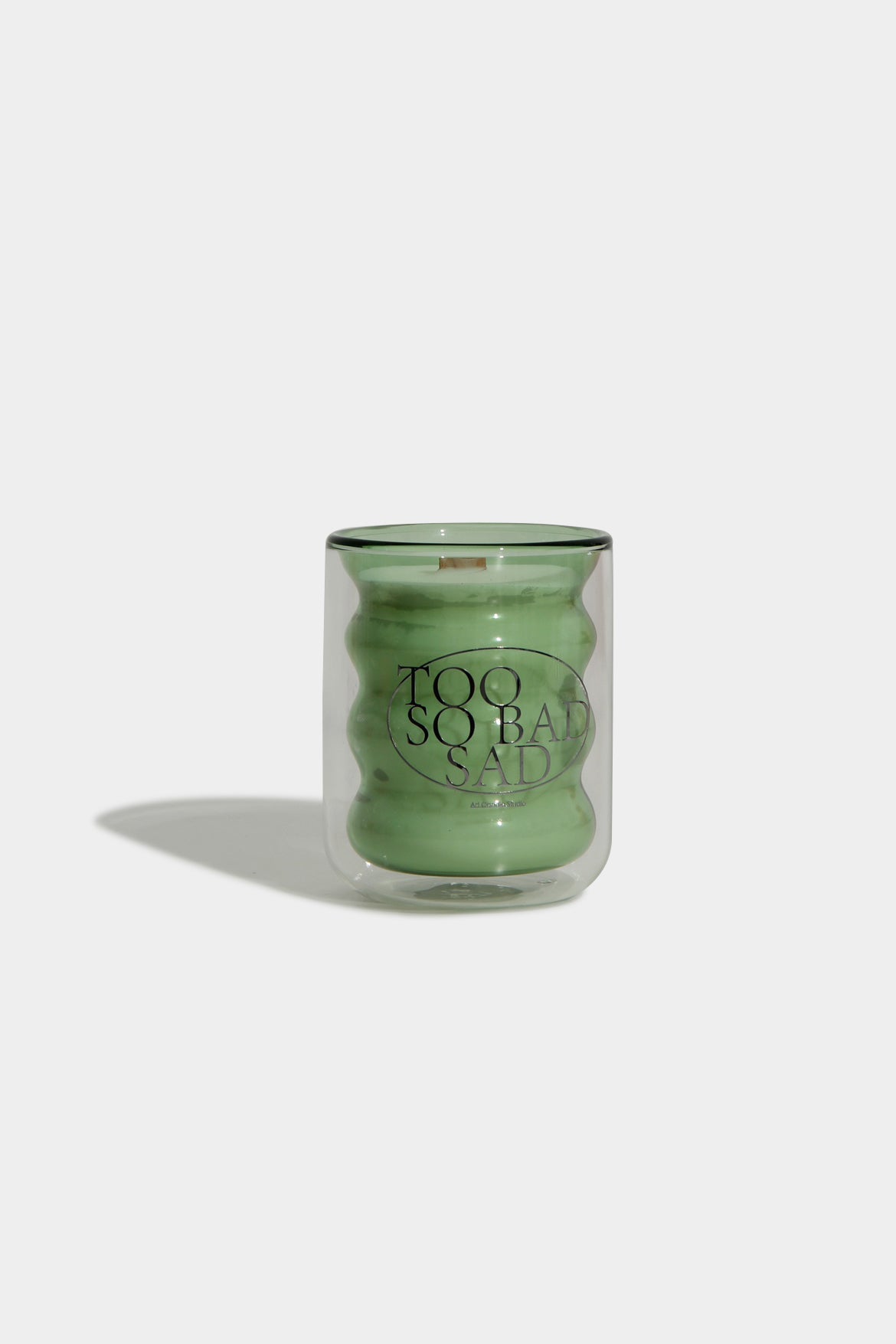 Green glass candle with 'Too So Bad Sad' text on a white background