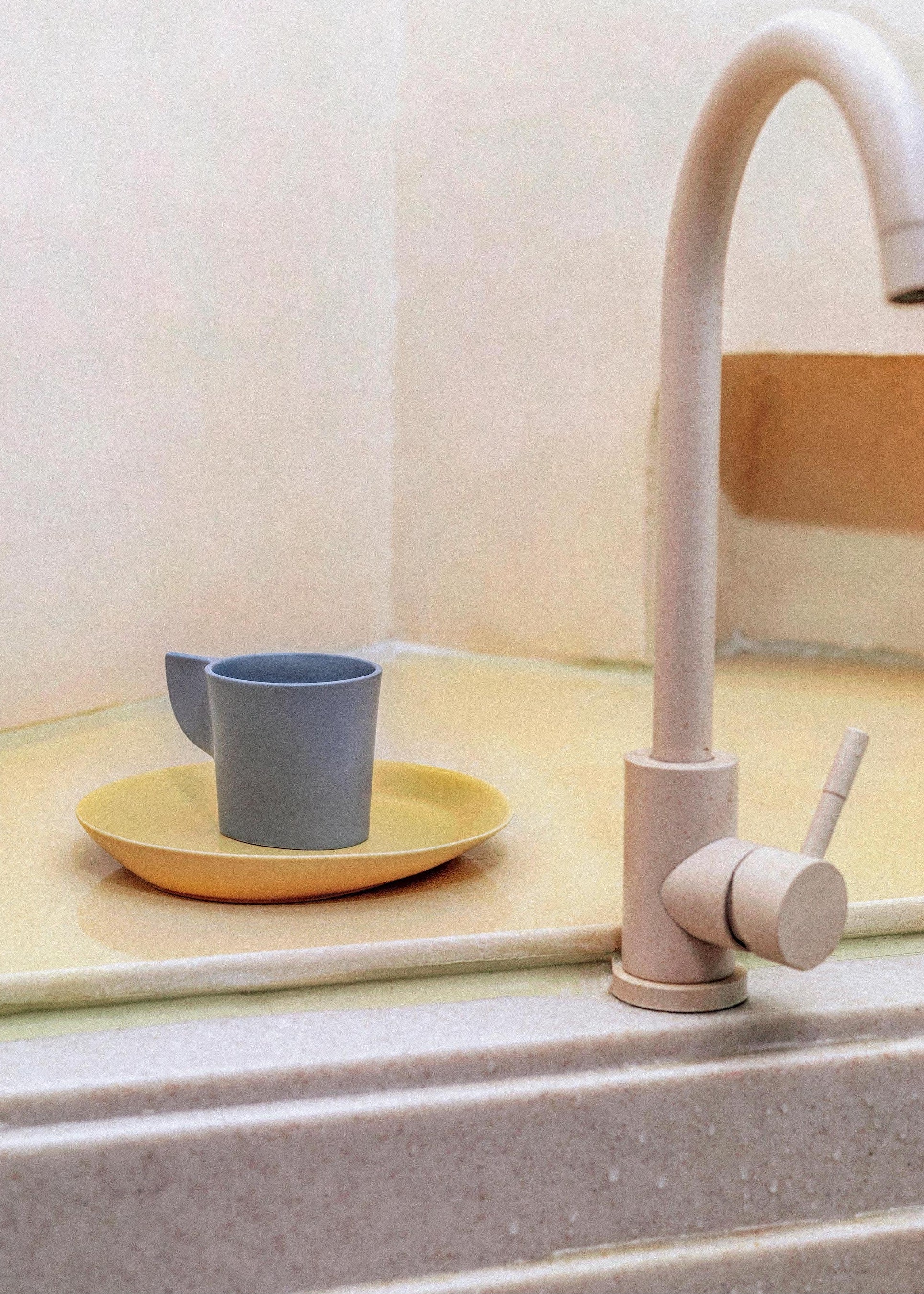 Kitchen sink with a beige faucet and a yellow plate with gray cups on a light-colored countertop.