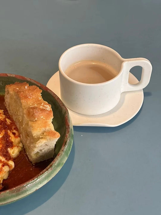 White ceramic cup with a handle on a saucer next to a plate of food on a gray surface