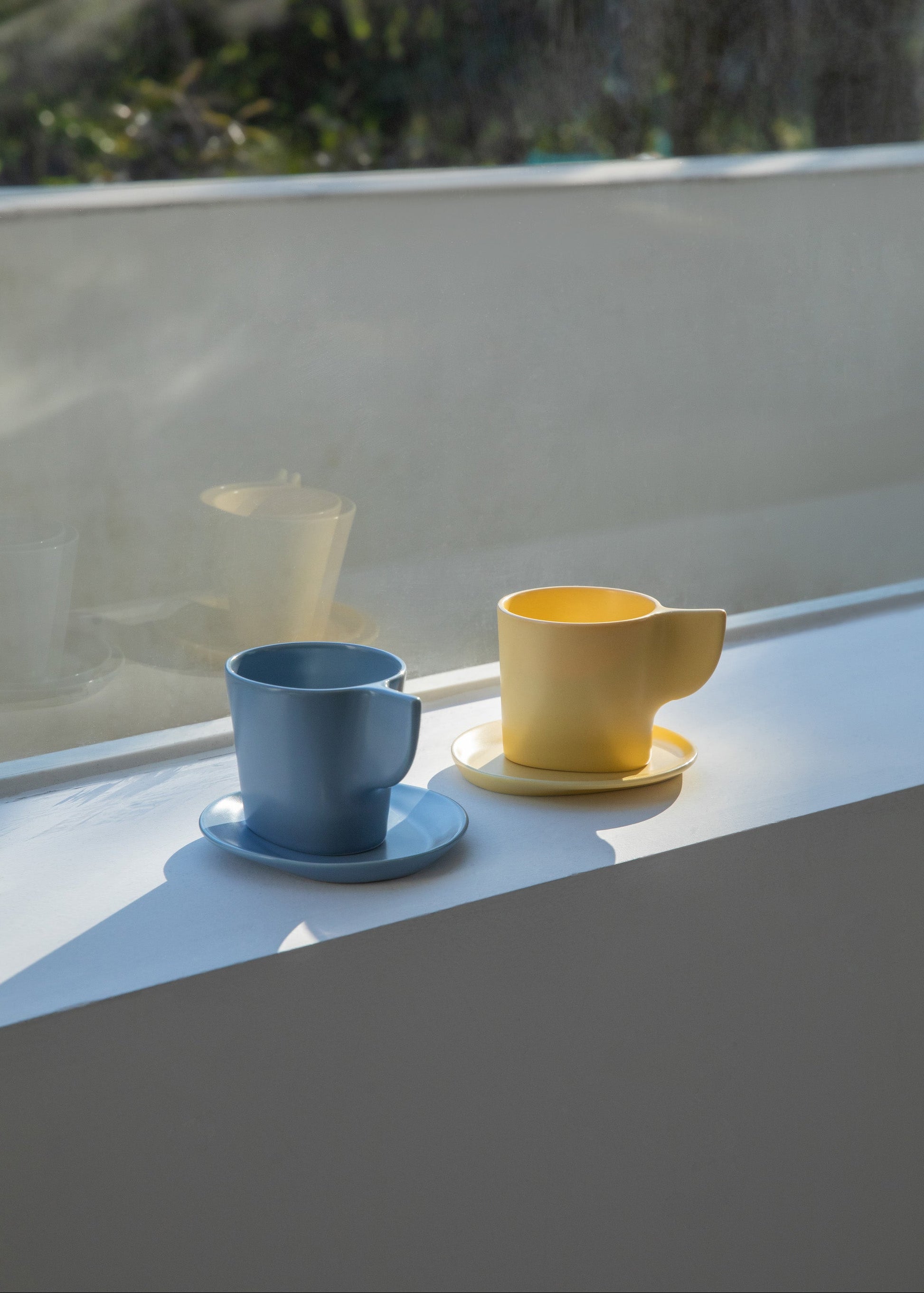Two ceramic cups, one blue and one yellow, on a reflective surface with a blurred natural background.