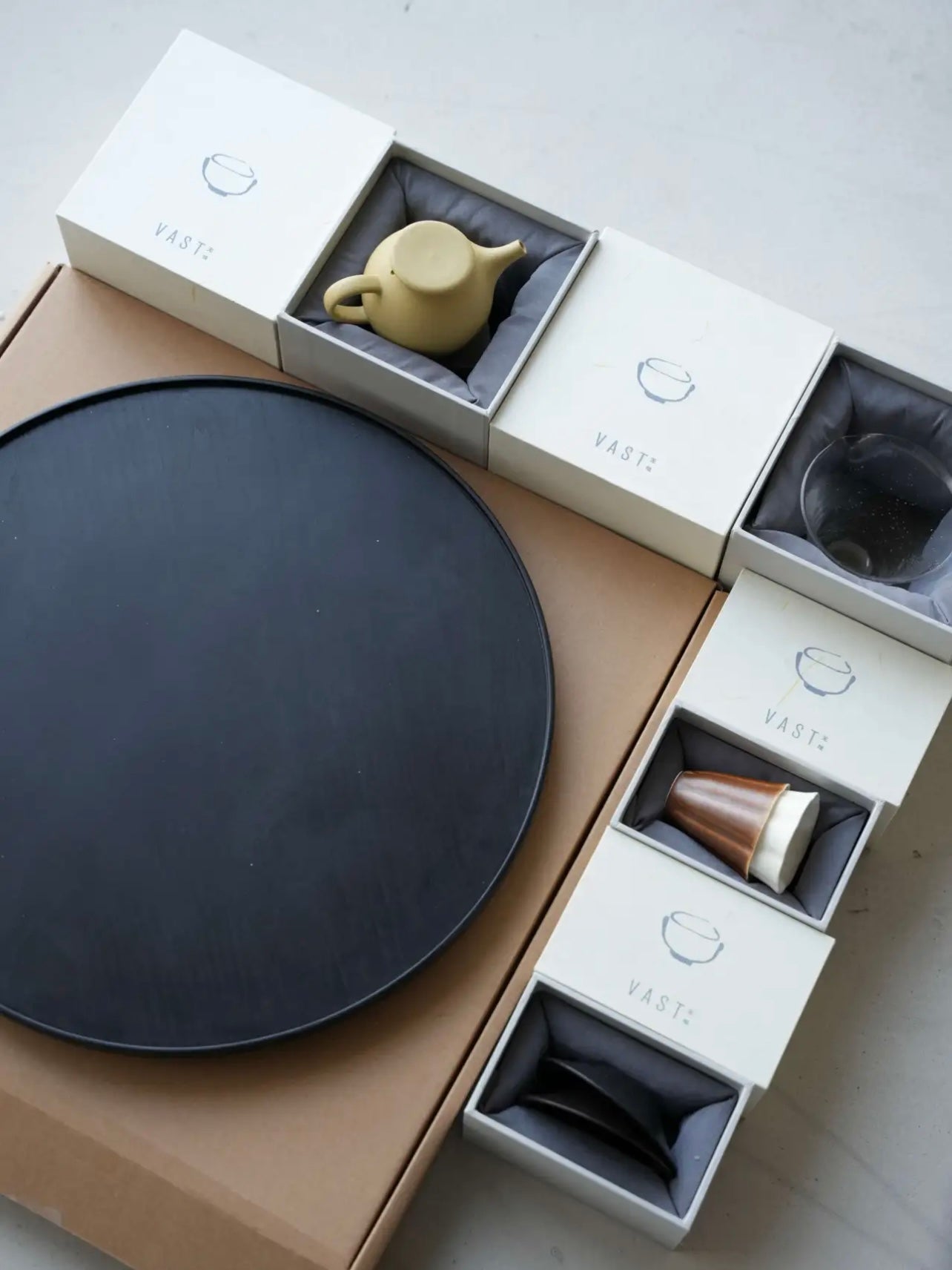 Set of tea-making items including a black round tray, teapot, and other accessories in open boxes on a light surface.