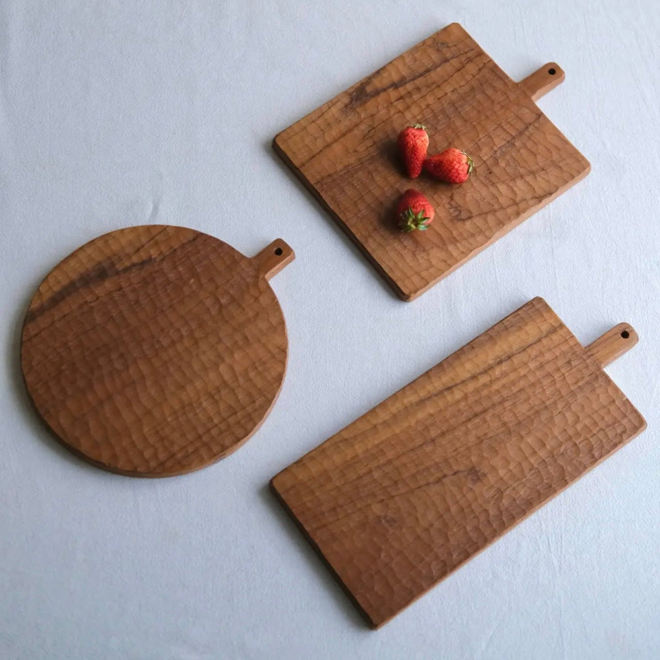 Three wooden cutting boards of different shapes on a white surface with strawberries on one of them.