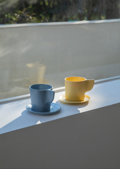 Two ceramic cups, one blue and one yellow, on a reflective surface with a blurred natural background.
